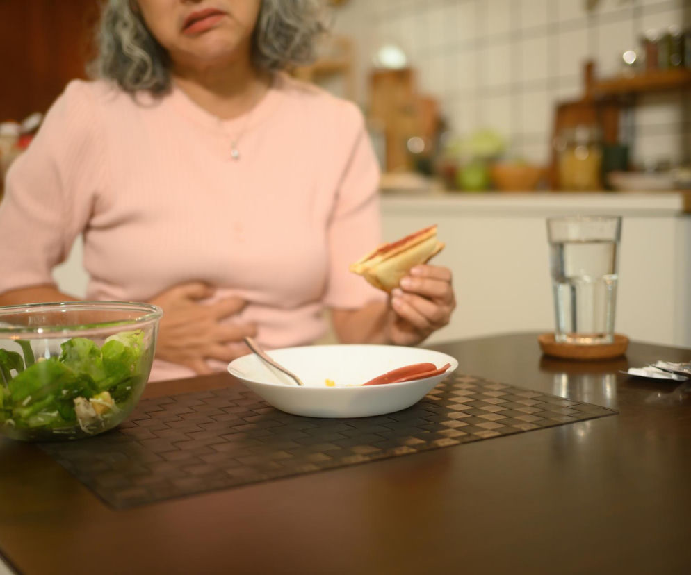 Starsza kobieta w jasnoróżowej bluzce, trzymająca kanapkę i dłoń na brzuchu, siedzi przy stole z sałatką, pustym talerzem, szklanką wody i lekami. Obraz symbolizuje problemy trawienne i poszukiwanie ulgi, o czym możesz przeczytać na Poradnik Zdrowie.