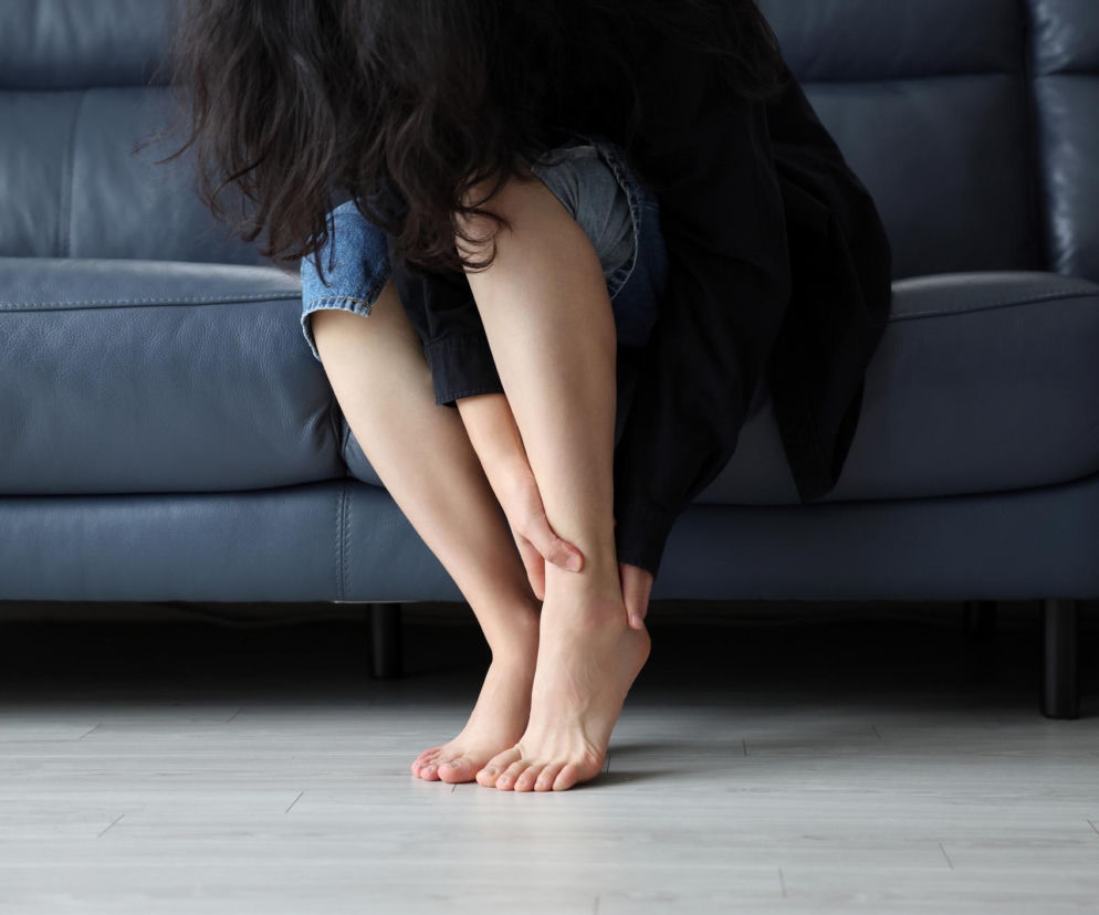 Kobieta siedząca na kanapie dotyka stopy i łydki, odczuwając mrowienie lub drętwienie nóg, co może być objawem wielu schorzeń. Na Poradnik Zdrowie znajdziesz informacje o przyczynach i leczeniu parestezji.