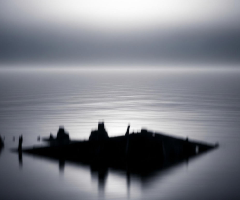 W centrum kadru, na powierzchni wody, widoczny jest ciemny, niewyraźny kształt przypominający wrak statku lub budowli, częściowo zanurzony, z fragmentarycznymi elementami wystającymi ponad lustro wody. Kształt ten rzuca odbicie na lekko pofalowaną, ciemną powierzchnię wody. Linia horyzontu jest wyraźna, oddzielając wodę od rozmytego nieba, które stopniowo jaśnieje od ciemniejszego błękitno-szarego odcienia przy horyzoncie, przechodząc w jaśniejszą, niemal białą poświatę w górnej części obrazu. Całość utrzymana jest w monochromatycznej palecie szarości i błękitów, z efektem rozmycia.