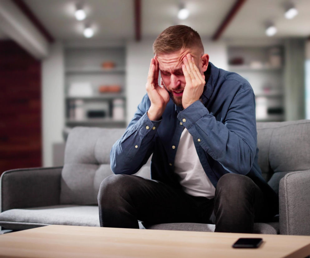 Mężczyzna w niebieskiej koszuli siedzi na kanapie, ściskając skronie w geście bólu głowy, objawu, który może wskazywać na problemy neurologiczne. Na Poradniku Zdrowie znajdziesz informacje o symptomach, które wymagają konsultacji lekarskiej.