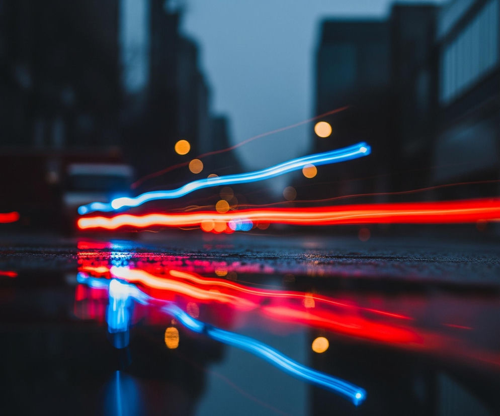 Ciemny, miejski krajobraz nocą, z rozmytymi smugami światła na pierwszym planie. Centralnie widoczne są intensywne, rozciągnięte smugi światła w kolorach czerwonym i niebieskim, biegnące poziomo przez dolną połowę kadru. Światła te, szczególnie te czerwone, odbijają się na mokrej, ciemnej powierzchni, tworząc dodatkowe, lekko zamazane odbicia. W tle znajdują się rozmyte kształty budynków w odcieniach szarości i granatu, a także kilka pojedynczych, pomarańczowych punktów świetlnych, przypominających lampy uliczne lub światła pojazdów w oddali.