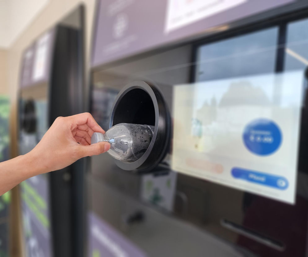 Dłoń osoby wkładającej plastikową butelkę PET do otworu automatu recyklingowego, na ekranie widoczne ikony interfejsu. Temat systemów kaucyjnych w Polsce i recyklingu opakowań omówiono na portalu Super Biznes.