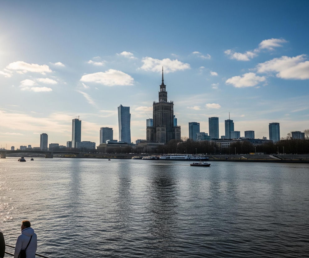 Połyskujące słońce, umiejscowione w lewym górnym rogu, oświetla szeroką rzekę, na której powierzchni widać liczne odbicia światła i małe fale. Na pierwszym planie, z prawej strony, dwóch mężczyzn w kurtkach stoi tyłem do obserwatora, opierając się o ciemną barierkę. W tle, na przeciwległym brzegu rzeki, rozciąga się miejska panorama z dominującym, wysokim budynkiem w centrum, otoczonym licznymi nowoczesnymi wieżowcami. Po rzece przemieszczają się mniejsze łodzie, a na horyzoncie widoczny jest most.