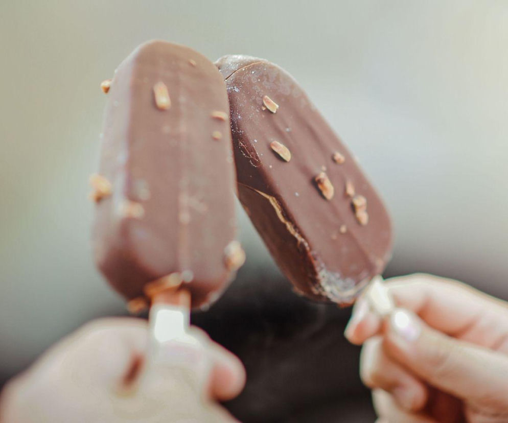 Dwie osoby trzymają lody na patyku w polewie czekoladowej z orzechami. O procesie produkcji lodów przeczytasz w Eskce.