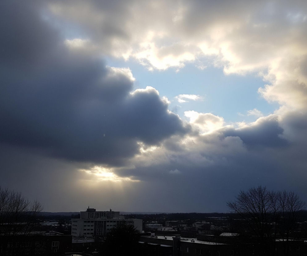 Niebo zdominowane jest przez ciemne, burzowe chmury, które tworzą nieregularny otwór w centrum, ukazujący fragment jasnego, błękitnego nieba. Promienie słońca przebijają się przez chmury w kilku miejscach, tworząc jasne plamy światła, szczególnie widoczne w dolnej części centralnego otworu. Poniżej rozpościera się ciemny, miejski krajobraz z widocznymi budynkami, w tym jednym większym, jasnym wieżowcem pośrodku. Na pierwszym planie po obu stronach kadru widać nagie drzewa o rozłożystych, ciemnych gałęziach, a w tle rozciągają się zarysy dalszych budynków i zalesionych wzgórz.