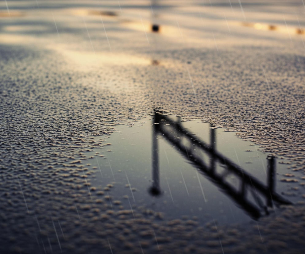 Na pierwszym planie widoczna jest mokra powierzchnia, pokryta licznymi drobnymi kroplami wody, które tworzą nieregularne wzory, przypominające pofałdowaną teksturę. Dominują ciemne odcienie szarości i brązu, z jaśniejszymi, złotawymi akcentami w górnej części kadru, sugerującymi odbicie światła. Po prawej stronie, w dolnej części obrazu, znajduje się kałuża z wyraźnym odbiciem ciemnego obiektu, przypominającego drabinę lub rusztowanie, ukośnie położonego w lustrzanej tafli wody. Całość jest skąpana w deszczu, co widać po licznym, równoległym białym liniom, przecinającym obraz pod kątem.