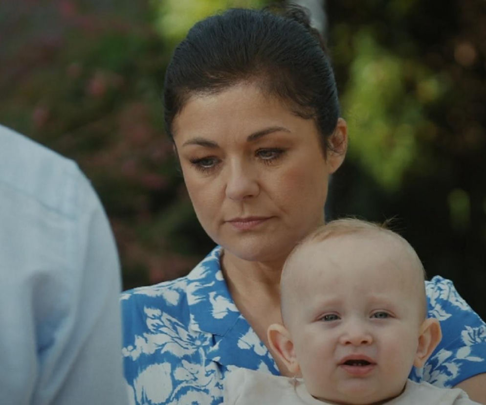 Katarzyna Cichopek (Kinga) w niebiesko-białej bluzce z kwiatowym wzorem trzyma na rękach niemowlę. Ma poważny wyraz twarzy, patrząc w dół. Fragment mężczyzny w jasnoniebieskiej koszuli jest widoczny po lewej stronie. W tle rozmazana zieleń. Więcej o losach bohaterów przeczytasz na Super Seriale.