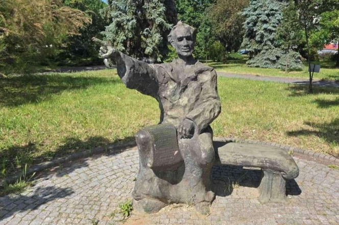 Rzeźba Jana z Głogowa siedzącego na ławce z księgą na kolanach i wskazującego prawą ręką w dal, umiejscowiona na brukowanej platformie w parku miejskim. Dookoła pomnika rozpościera się zielona trawa i bujna roślinność, w tle widoczne wysokie drzewa. O dewastacji pomnika przeczytasz więcej na naszym portalu.