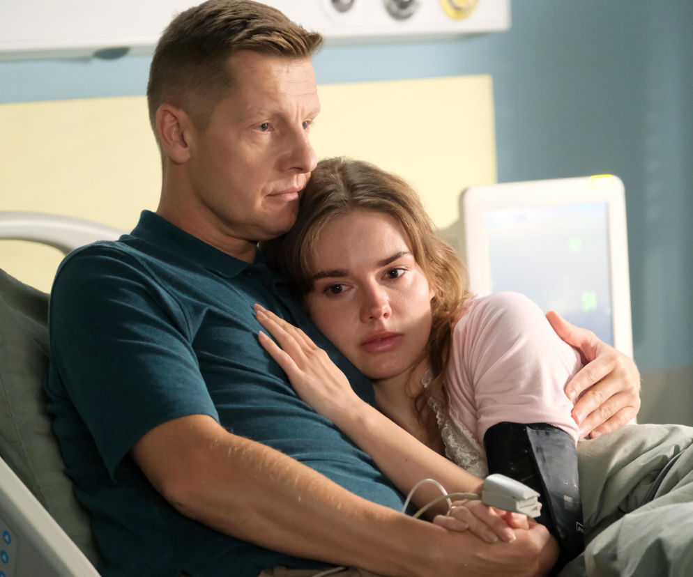 Paweł Mroczek obejmujący Dominikę Kachlik w szpitalnym łóżku, z monitorem medycznym w tle. Kobieta ma na sobie różową koszulkę, a on zieloną koszulkę polo. Ujęcie odzwierciedla smutek i wsparcie w trudnym momencie odejścia Franki z serialu, o czym więcej przeczytasz na stronie Super Seriale.