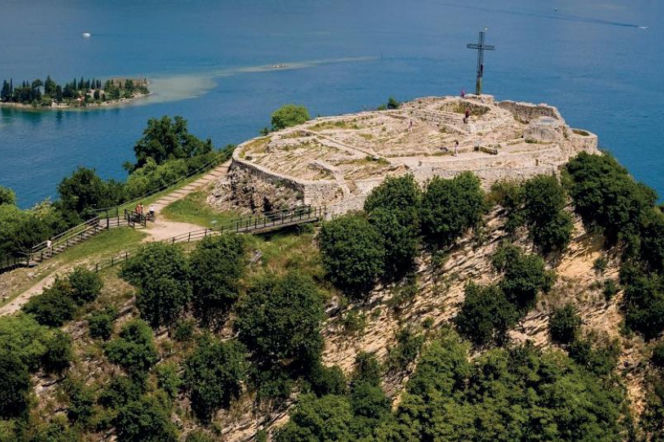 Widok z lotu ptaka na Rocca di Manerba, historyczną twierdzę na skalistym cyplu z metalowym krzyżem, otoczoną bujną roślinnością. W tle błękitne wody jeziora Garda i wyspa San Biagio. Odkryj sekrety tego miejsca na naszym portalu.