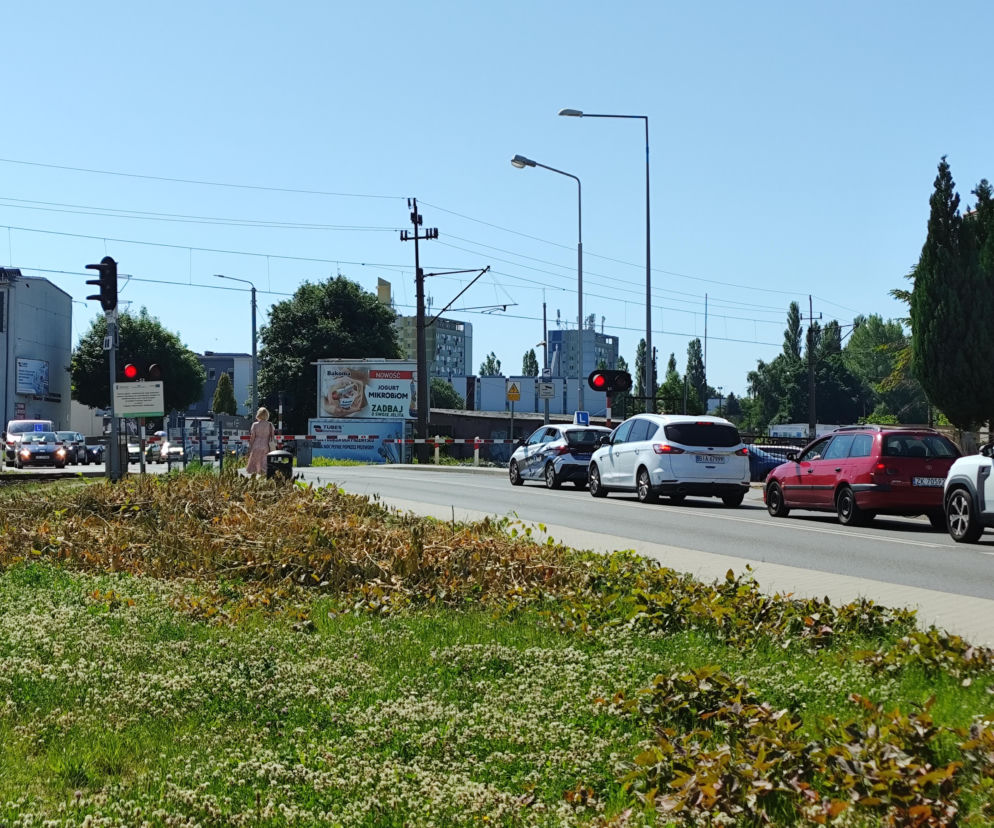 Ulica Franciszkańska w Koszalinie z przejazdem kolejowym i zamkniętymi rogatkami, gdzie widoczne są stojące samochody, sygnalizator z czerwonym światłem oraz kobieta, która spaceruje obok. Kontekst bezpiecznego przejazdu kolejowego dostępny jest na naszym portalu.