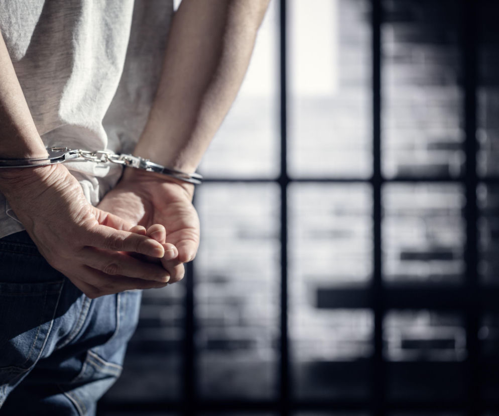 Dłonie mężczyzny zakute w kajdanki na tle więziennych krat. O wyroku 17 lat więzienia za pobicie przeczytasz na SE.
