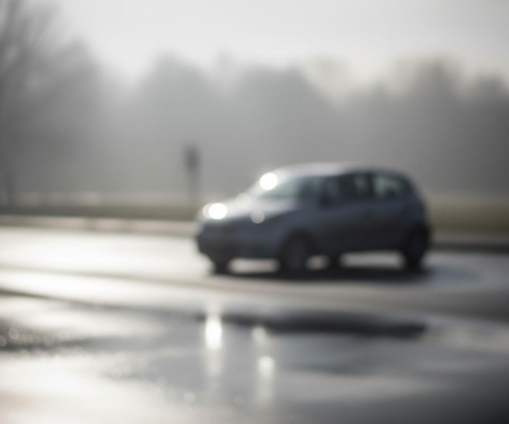 Rozmyte zdjęcie przedstawia samochód osobowy, prawdopodobnie szarej lub srebrnej barwy, poruszający się po drodze. Na pierwszym planie widać mokrą nawierzchnię, z refleksami światła odbijającymi się od kałuż, tworzącymi efekt bokeh. W tle rysują się niewyraźne sylwetki drzew lub zarośli, spowite mgłą lub dymem, nadając kompozycji tajemniczy i rozmyty charakter.