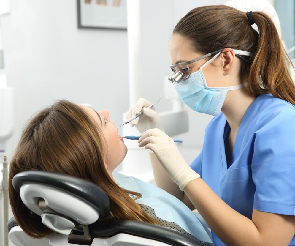 Dentystka w masce, okularach i jednorazowych rękawiczkach podczas przeglądu jamy ustnej pacjentki leżącej na fotelu. Zadbaj o higienę, aby zapobiegać chorobom serca – więcej na ten temat przeczytasz na portalu Poradnik Zdrowie.