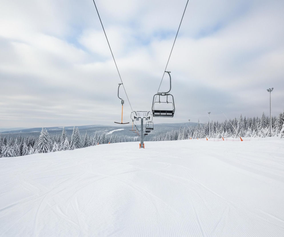 Wyciąg krzesełkowy biegnie przez środek szerokiej, białej, pokrytej śniegiem trasy narciarskiej, która opada w dół od prawego górnego rogu. Trasa jest świeżo naśnieżona, z wyraźnymi równoległymi śladami ratraka oraz kilkoma nieregularnymi śladami nart. Po prawej stronie widoczne są pomarańczowe siatki zabezpieczające i jeden pomarańczowy, przysypany śniegiem pachołek. W tle, po obu stronach trasy, rozciąga się gęsty, zaśnieżony las iglasty, a dalej widoczny jest odległy horyzont. Niebo jest pochmurne, z wyraźnymi smugami szarych i błękitnych chmur.