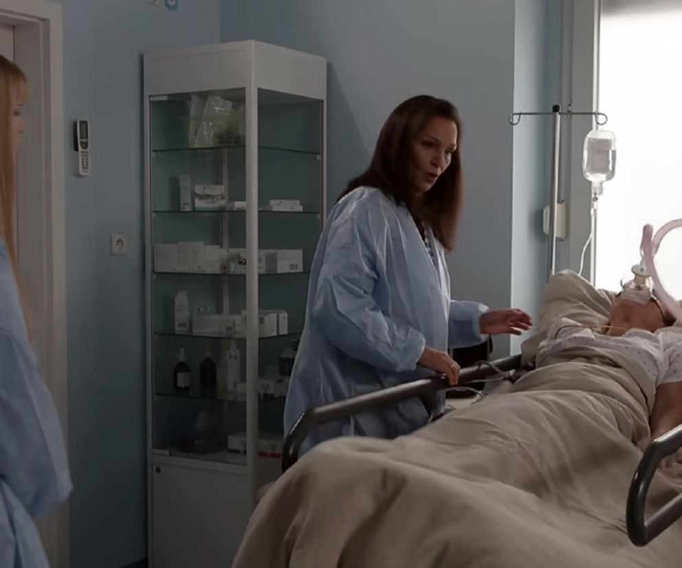 Scena w szpitalu z serialu Pierwsza miłość, gdzie Marysia (Aneta Zając) leży nieprzytomna w łóżku z maską tlenową, a nad nią pochyla się Izabela Nowaczyk (Olga Bończyk) w jednorazowym fartuchu medycznym. Obok stoi Julka (Dąbrówka Kruk). Więcej o dramatycznych wydarzeniach w Super Seriale.