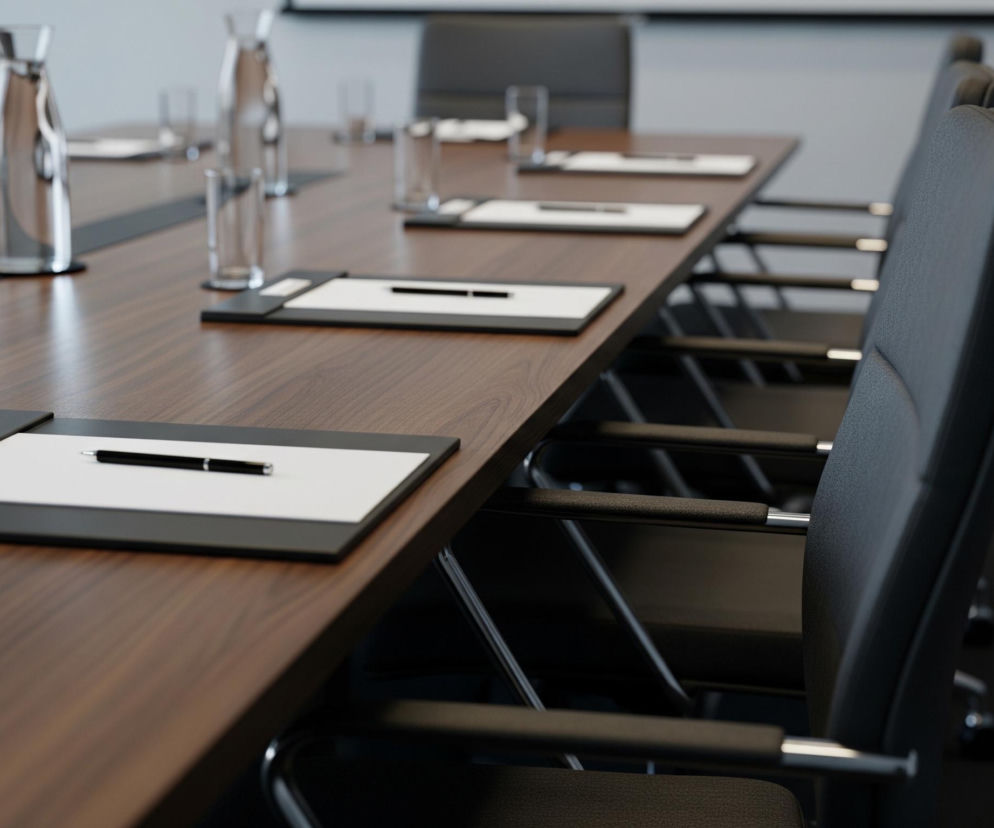 Centralne miejsce zajmuje długa, drewniana deska stołu o brązowym, błyszczącym wykończeniu, rozciągająca się od lewego dolnego rogu w głąb obrazu. Na stole, w regularnych odstępach, leżą czarne podkładki z białymi kartkami papieru i czarnymi długopisami, a obok nich stoją puste szklanki i szklane karafki z wodą. Wzdłuż prawej strony stołu ustawione są rzędy czarnych, skórzanych krzeseł z metalowymi podłokietnikami, skierowanych w stronę stołu. Tło stanowi jasnoszara ściana oraz ciemna podłoga, a w oddali widać biały ekran projektora.