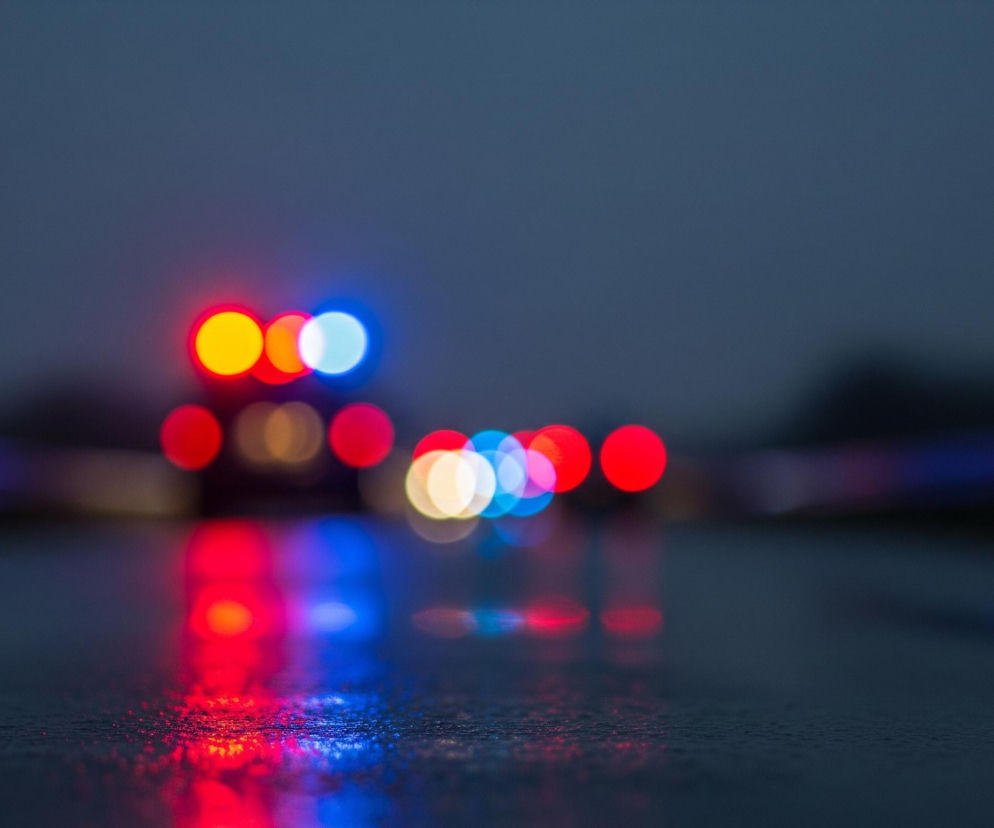 Rozmyte światła w kolorach czerwonym, pomarańczowym, białym i niebieskim dominują centralną część obrazu. Światła te odbijają się na mokrej, ciemnej powierzchni, tworząc jaskrawe, rozmazane refleksy. Tło jest ciemne i niewyraźne, z delikatnymi zarysami kształtów, sugerującymi odległe obiekty lub otoczenie miejskie.
