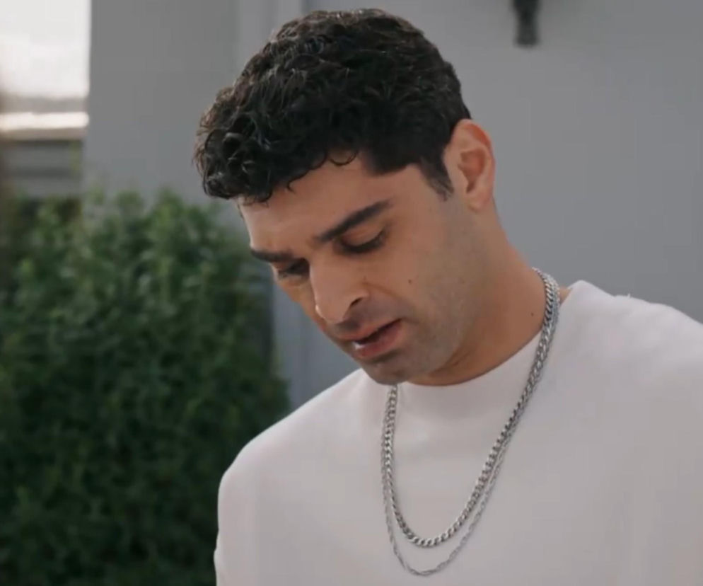 Mężczyzna o ciemnych, kręconych włosach i śniadej cerze, ubrany w białą bluzę i dwa srebrne łańcuszki, patrzy w dół z wyraźnym zamyśleniem. Postać symbolizuje emocje związane z fabułą serialu Złoty chłopak, o czym więcej można przeczytać na portalu Super Seriale.