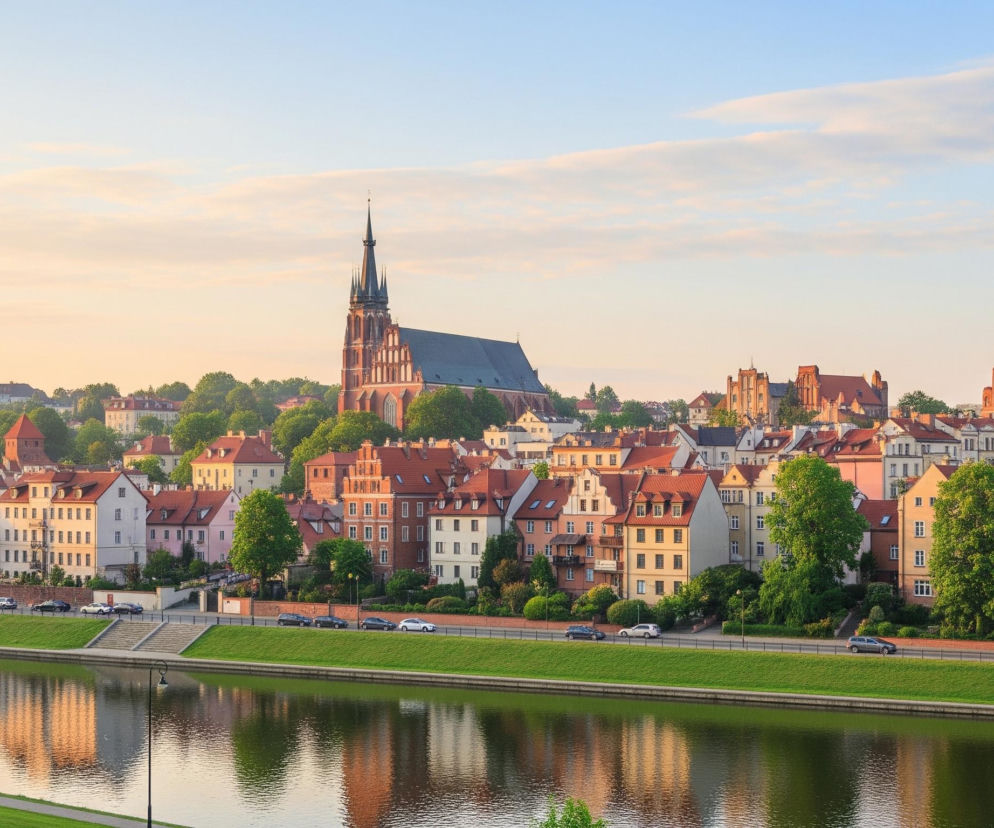 Szeroki kadr ukazuje miasto położone nad rzeką w świetle wczesnego poranka lub późnego popołudnia, z niebieskim niebem przechodzącym w jaśniejsze odcienie żółci i pomarańczu przy horyzoncie. Dominującym elementem panoramy jest gotycki kościół z wysoką, strzelistą wieżą i ciemnym dachem, otoczony gęstą zielenią drzew. Poniżej rozciąga się rzędowa zabudowa z ceglanymi lub pastelowymi fasadami i czerwonymi dachami, odzwierciedlającymi się w spokojnej tafli rzeki. Wzdłuż rzeki biegnie ulica z kilkoma zaparkowanymi samochodami, oddzielona od wody wąskim pasem zielonej trawy i schodami.