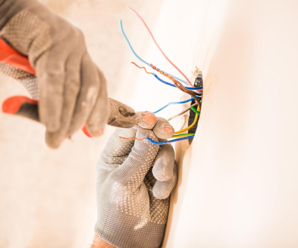 Ręce elektryka obcinające kable przy puszce w ścianie. O cenach instalacji w 2026 roku przeczytasz na Muratordom.