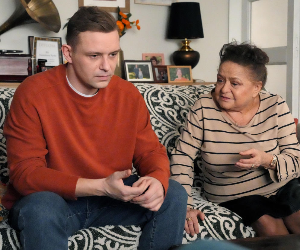 Hubert (Marek Molak) w pomarańczowym swetrze siedzi na kanapie z wzorzystym kocem, obok Lucyna (Grażyna Zielińska) w beżowej bluzce w paski i czarnej spódnicy. Oboje wyglądają na zmartwionych, gdy Lucyna coś opowiada Hubertowi. To fragment Barw Szczęścia, o którym przeczytasz więcej na Super Seriale.