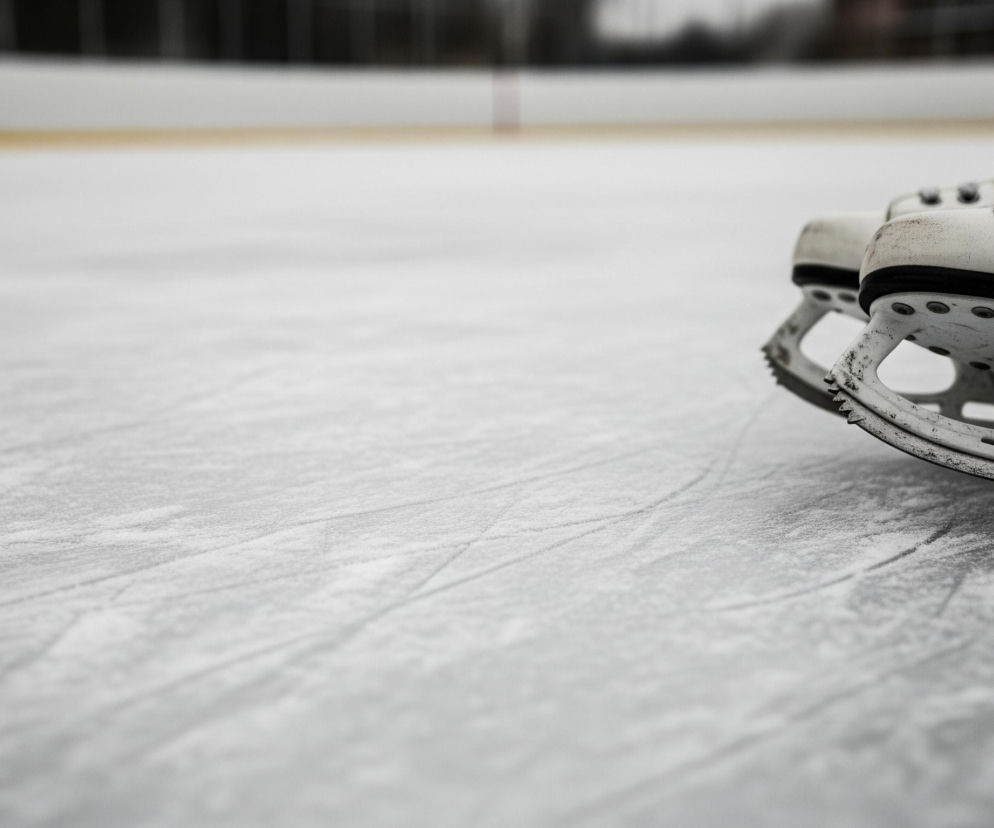 Po prawej stronie zdjęcia leżą dwie białe łyżwy figurowe na tafli lodu. Są one ułożone równolegle do siebie, jedna za drugą, z ostrzami skierowanymi w lewo. Na powierzchni lodu widoczne są ślady ostrzy łyżew, a w tle zarys barierki otaczającej lodowisko.