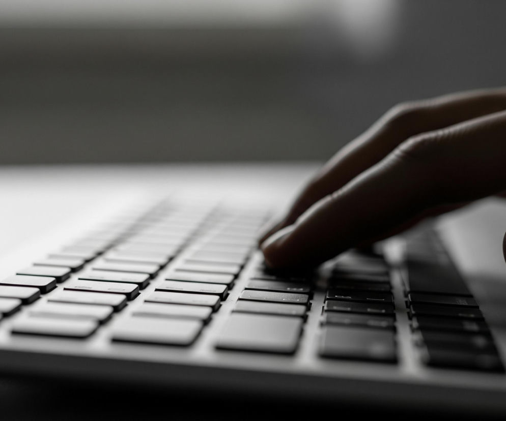 Sylwetka prawej dłoni widoczna jest w górnej, prawej części zdjęcia, jej palce zginają się nad klawiaturą laptopa, dominują tu ciemne odcienie. Klawisze laptopa, ułożone w rzędy, są przeważnie białe lub jasnoszare, jedynie te najbliżej dłoni są ciemne, co sugeruje odbicie światła. Reszta laptopa to jasna powierzchnia, rozświetlona mocno po lewej stronie. Tło jest rozmyte, utrzymane w szaro-białej tonacji.