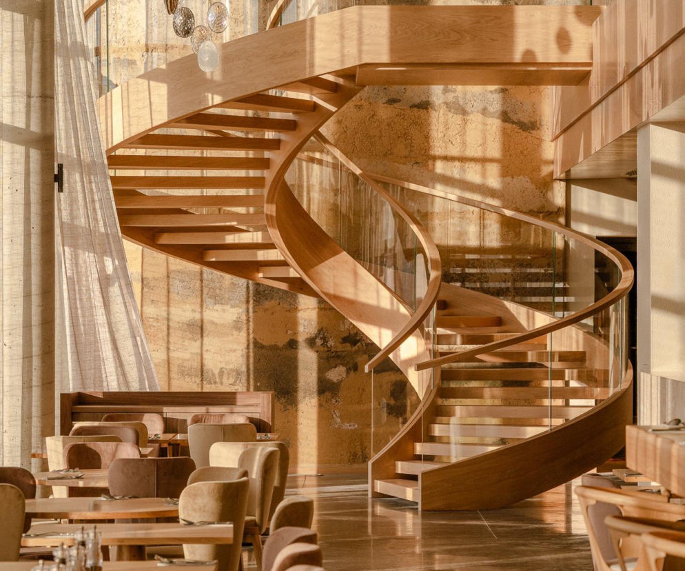 Drewniana spiralna klatka schodowa z przeszklonymi barierkami w lobby Hotelu GLAR, którego projekt autorstwa Piotra Maciaszka i Karola Pasternaka można poznać dzięki Architektura Murator Plus. Obok minimalistyczne stoły z krzesłami, za nimi ściana z tynku glinianego.