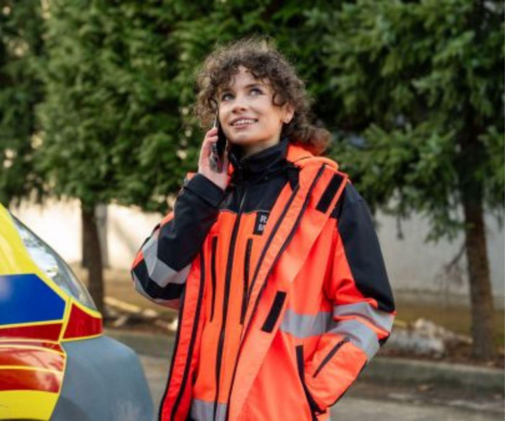 Uśmiechnięta Martyna w pomarańczowej kurtce ratownika medycznego rozmawia przez telefon, stojąc obok karetki, co odzwierciedla codzienne wyzwania bohaterów serialu „Na sygnale”. Więcej o ich perypetiach można przeczytać na Super Seriale.