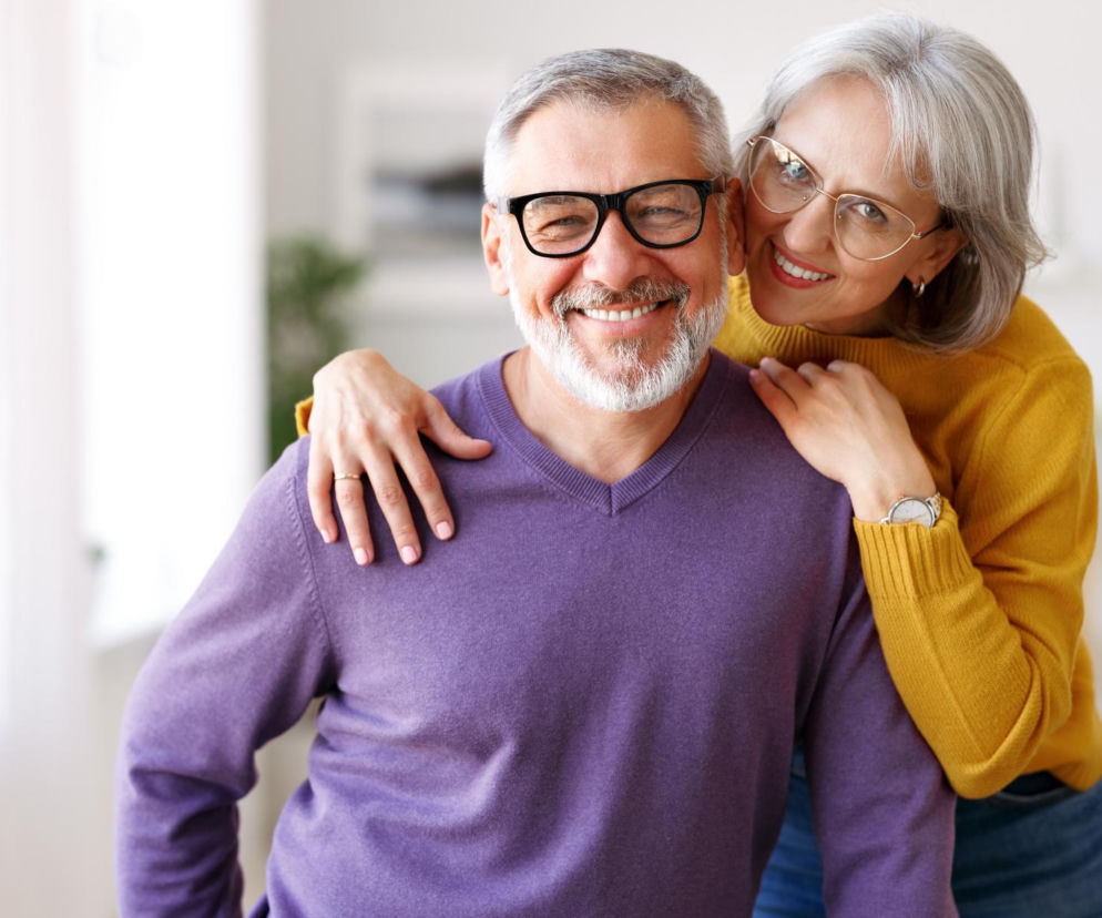 Szczęśliwe małżeństwo emerytów, mężczyzna w fioletowym swetrze i okulary, kobieta w żółtym swetrze, uśmiechają się, stojąc blisko siebie, co symbolizuje oszczędności i zysk dla seniorów. Więcej o zwrocie podatku dla małżeństw emerytów przeczytasz na Super Biznes.