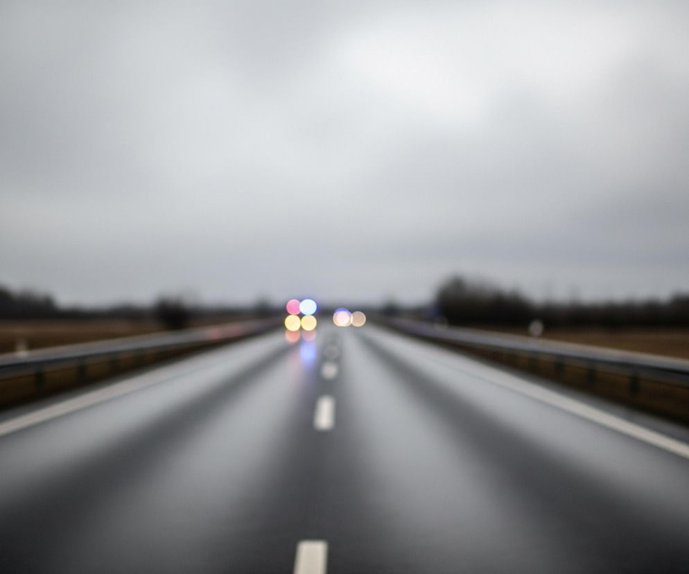 Widoczna jest droga asfaltowa, mokra i ciemnoszara, rozciągająca się od dołu obrazu w kierunku centrum, z białą przerywaną linią pośrodku. Po obu stronach drogi biegną metalowe barierki ochronne. Tło stanowi rozmyty krajobraz z ciemnymi drzewami i brązowymi polami, a nad nim ciężkie, szare niebo. W oddali na drodze widać rozmyte, kolorowe światła samochodów: czerwone, niebieskie i żółte.