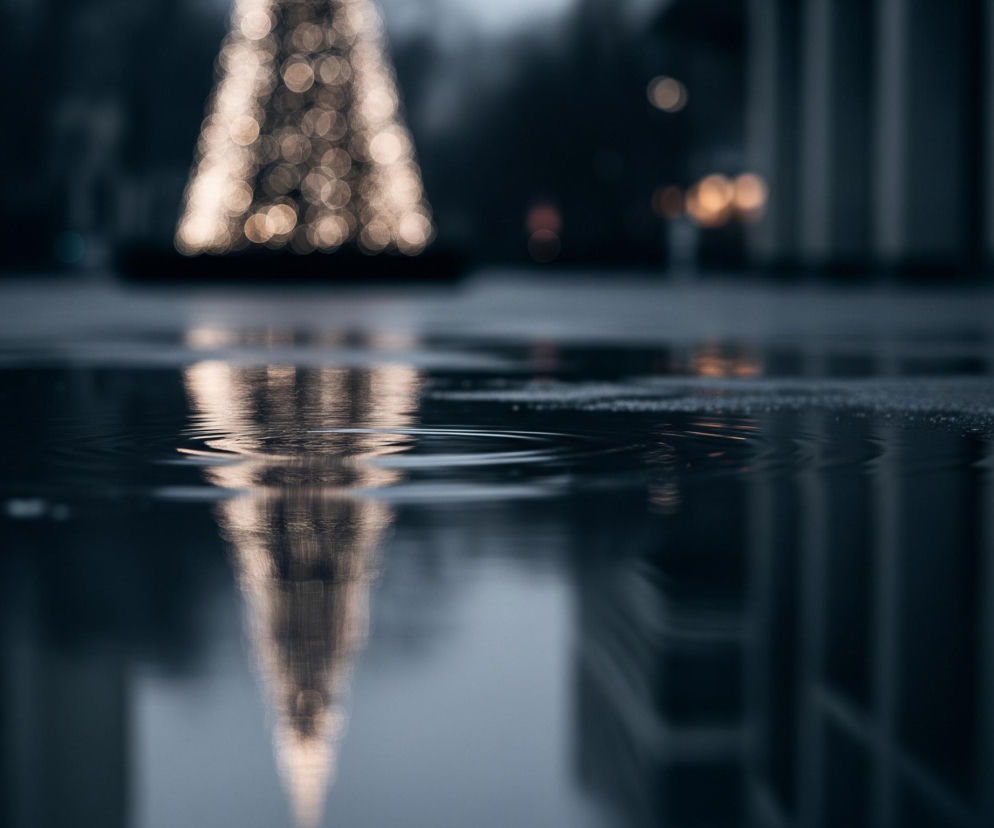 Głównym elementem jest rozmyte odbicie iluminacji świetlnej w kształcie choinki, widoczne w kałuży na pierwszym planie, z delikatnymi falami na powierzchni wody. W tle, powyżej kałuży, znajduje się rzeczywista, niewyraźna choinka utworzona ze złocistych, okrągłych światełek typu bokeh. Po prawej stronie widać rozmyte, pionowe elementy architektoniczne budynków oraz dalsze, niewyraźne światła. Dominują ciemne, chłodne barwy niebieskiego i szarego, przełamane ciepłym, rozproszonym światłem.