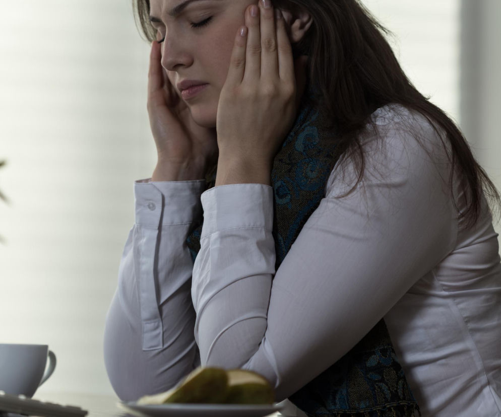 Kobieta z zamkniętymi oczami i dłońmi przy skroniach, sprawiająca wrażenie zdenerwowanej lub zmęczonej, z szalikiem na szyi, siedząca przy biurku z filiżanką i talerzykiem. Temat mizofonii i jej objawów znajdziesz na Poradnik Zdrowie.