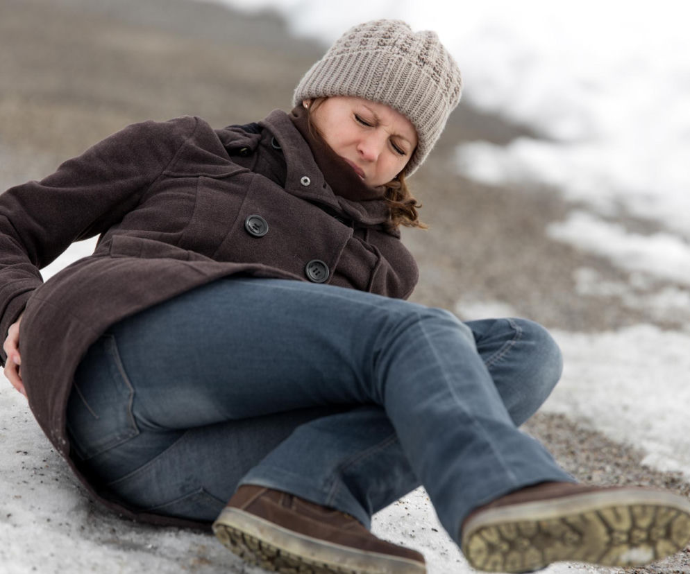 Kobieta w brązowym płaszczu i zimowej czapce siedzi na oblodzonym chodniku, trzymając się za plecy po upadku. Wyraża ból i dyskomfort. Dowiedz się, jak unikać kontuzji zimą i co robić po poślizgnięciu, czytając na Poradnik Zdrowie.