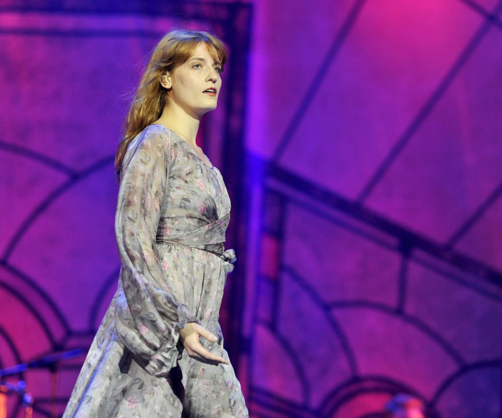 Florence Welch w zwiewnej, kwiecistej sukience podczas występu na scenie, z długimi, rudymi włosami, na fioletowym tle. Więcej szczegółów o występie zespołu Florence + the Machine na Open'er Festival 2026 znajdziesz na naszym portalu.