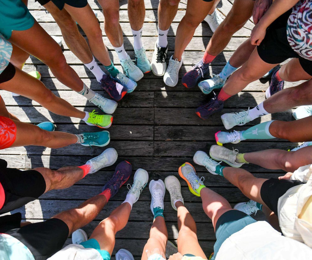 Grupa osób w różnokolorowych butach do biegania marki ASICS, ułożonych w okrąg na drewnianej podłodze, symbolizująca aktywność fizyczną i sukces marki. O wynikach finansowych ASICS przeczytasz na Super Biznes.