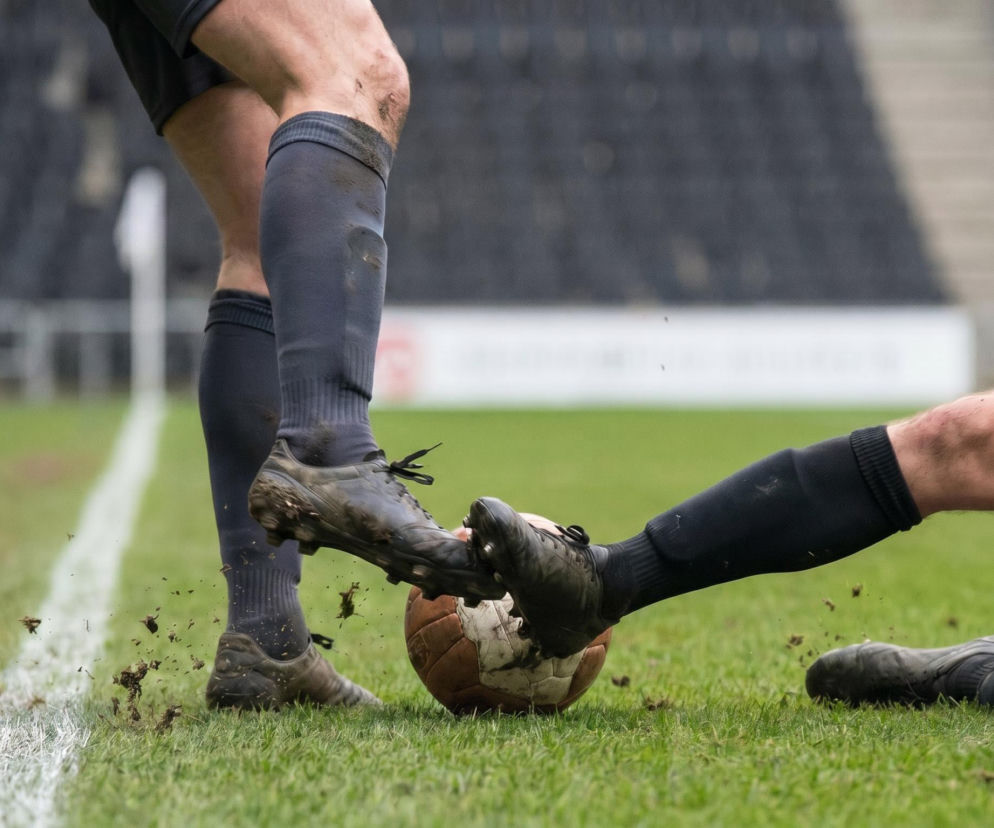 Nogi piłkarzy w ciemnych strojach walczą o brudną, starą piłkę na zielonej murawie, blisko białej linii boiska. W tle widać puste trybuny stadionu, co nawiązuje do relacji z meczu Kolumbia - Portugalia na naszym portalu.