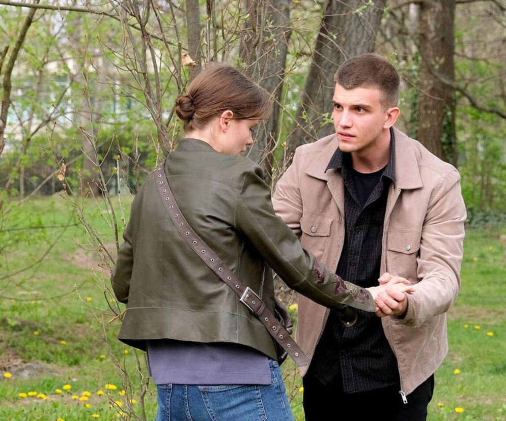 Mateusz (Rafał Kowalski) i Majka (Matylda Giegżno) z serialu M jak miłość na tle zielonego parku. Mateusz trzyma Majkę za rękę, spoglądając na nią z zatroskaniem, podczas gdy dziewczyna odwraca się plecami, ubrana w zieloną kurtkę i dżinsy. Artykuły o ich losach znajdziesz na Super Seriale.