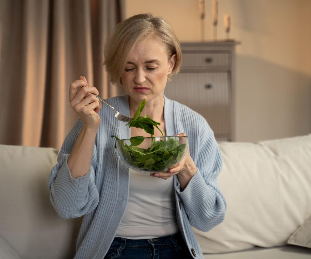 Kobieta w średnim wieku z krótkimi, blond włosami, siedząca na beżowej sofie, z niechęcią patrzy na widelec z zieloną sałatą, trzymając szklaną miskę. Jej mina wskazuje na nieprzyjemne odczucia smakowe, co ma związek z utratą smaku, o której można przeczytać na Poradnik Zdrowie.