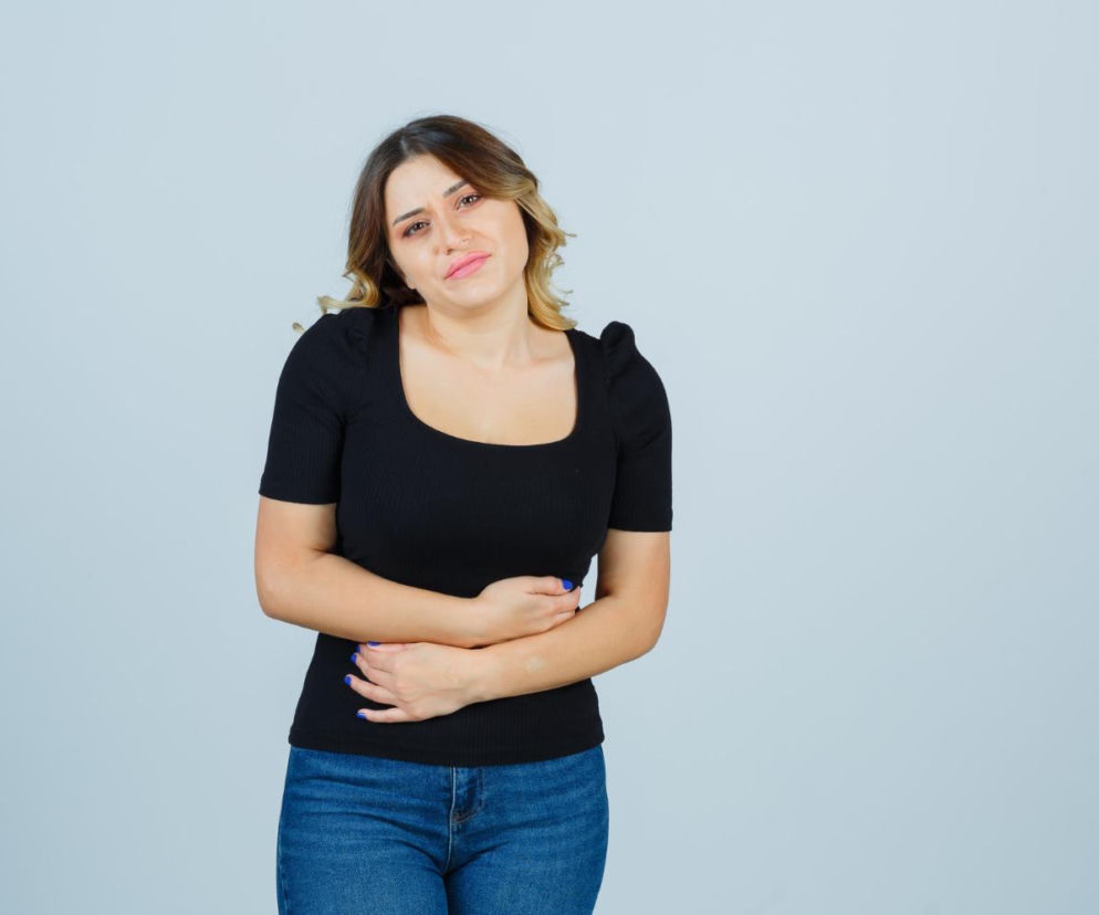 Kobieta w czarnym topie i jeansach trzyma się za brzuch, sygnalizując dyskomfort związany z pulsowaniem w jamie brzusznej. Na Poradnik Zdrowie znajdziesz informacje, kiedy pulsowanie jest groźne i może wskazywać na tętniaka aorty.