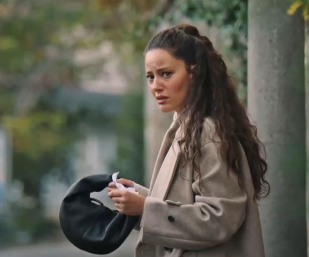 Diyar ze serialu Złoty chłopak, z zaniepokojoną miną trzymająca chusteczkę i torebkę, ubrana w beżowy płaszcz, przedstawiona na tle zieleni. Więcej o losach bohaterki na portalu Super Seriale.