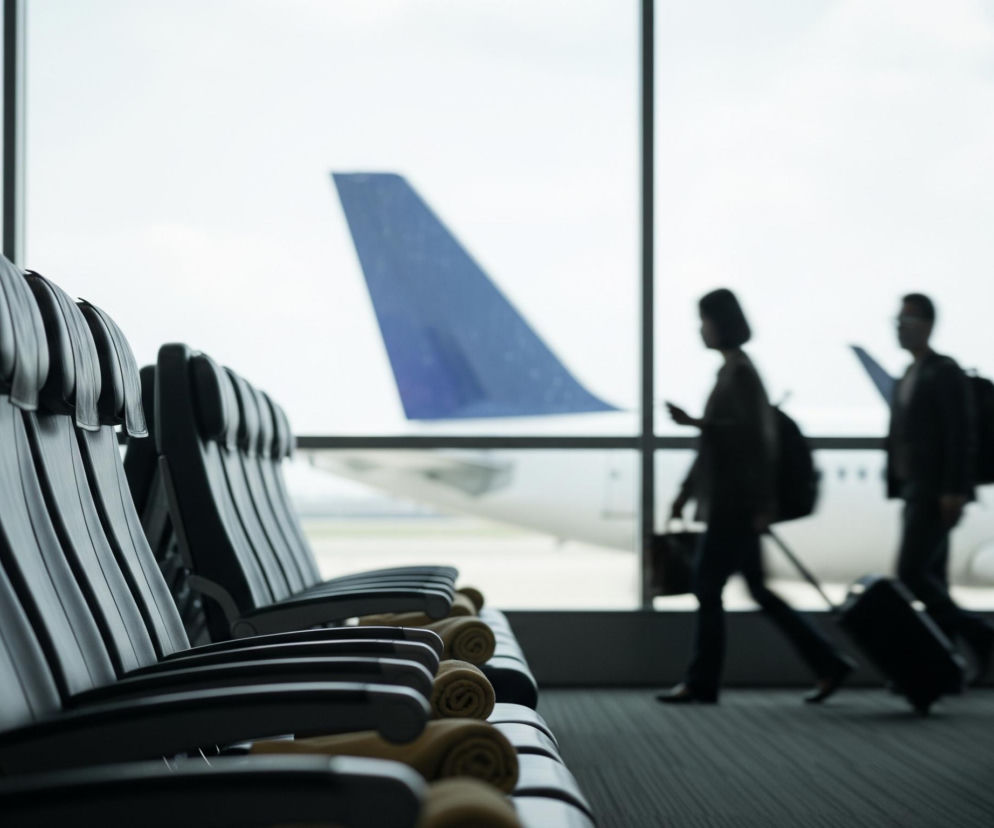 Rząd ciemnych, pustych foteli rozciąga się w kierunku prawej strony zdjęcia, tworząc perspektywiczną linię. Za fotelami, przez duże okno, widoczna jest sylwetka samolotu z niebieskim, pionowym statecznikiem. Trzy rozmyte postacie, ciemne sylwetki na tle jasnego nieba, idą w prawo, niosąc bagaże podręczne, w tym jedną walizkę na kółkach i plecaki.