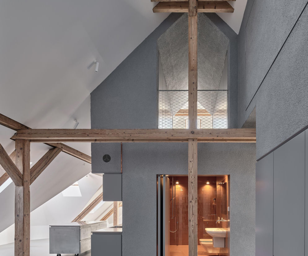 Jasne poddasze kamienicy w Gliwicach z drewnianą więźbą dachową i nowoczesnym aneksem kuchennym w szarościach, tworzące harmonijną przestrzeń. Wyróżnia się łazienka z miedzianymi kafelkami. O projekcie pracowni Dyrda Fikus Architekci przeczytasz na Architektura Murator Plus.