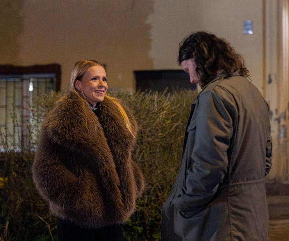 Aneta Zając jako Dominika, ubrana w futrzane brązowe okrycie, uśmiecha się do Mateusza Kościukiewicza jako Pawła Krzyżanowskiego w ciemnej kurtce. Scena rozgrywa się wieczorem przed budynkiem, w 4207 odcinku Pierwszej miłości, o którym przeczytasz więcej na Super Seriale.