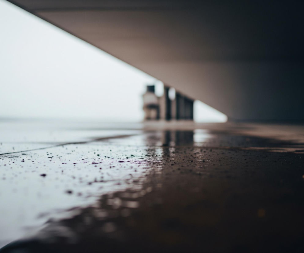 Mokra, ciemnoszara nawierzchnia pokryta jest licznymi kroplami wody i smugami, co nadaje jej błyszczący wygląd. W lewej części podłoża widać kilka drobnych, rozrzuconych kropek o zielonkawym i fioletowym zabarwieniu. Horyzont na zdjęciu jest rozmyty, a nad nim rozciąga się jasne, szare niebo, które przechodzi w ciemniejszy, ukośny fragment architektoniczny w górnej części kadru. Z prawej strony, pod ukośnym elementem, wyłaniają się niewyraźne, pionowe kształty budowli.