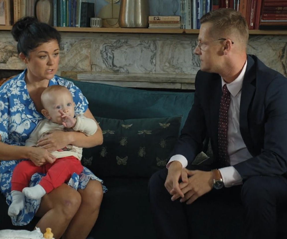 Kinga Zduńska (Katarzyna Cichopek) z Antosiem na rękach, rozmawia z Piotrkiem Zduńskim (Marcin Mroczek) w salonie. Kinga ma na sobie niebieską sukienkę w białe kwiaty, Antoś w kremowej bluzce i czerwonych spodniach. Oboje siedzą na granatowej kanapie, a obok nich leży torba z akcesoriami dziecięcymi. Więcej informacji o serialu M jak miłość znajdziesz na Super Seriale.