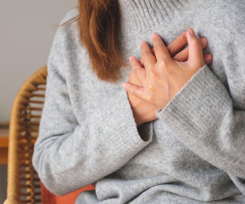 Kobieta w szarym swetrze trzymająca się za klatkę piersiową, symbolizując kołatanie serca lub ból, co jest tematem artykułu o momentach, w których trzeba reagować. Więcej o kołataniu serca przeczytasz na Poradnik Zdrowie.