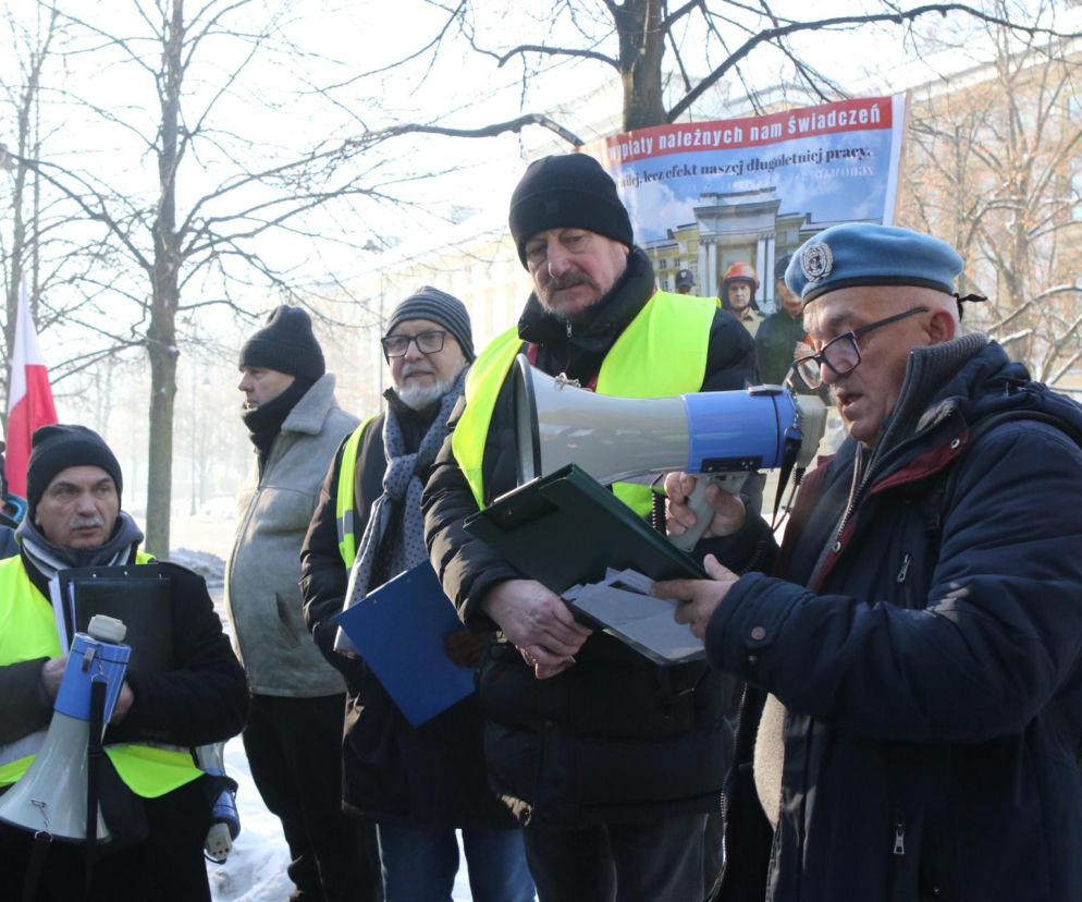 Grupa emerytowanych funkcjonariuszy mundurowych, ubranych w żółte kamizelki i zimowe czapki, podczas protestu pod Kancelarią Premiera. Jeden z nich przemawia przez megafon, trzymając dokumenty. Transparent w tle głosi: „Wypłaty należnych nam świadczeń”. Więcej na ten temat przeczytasz na Super Biznes.