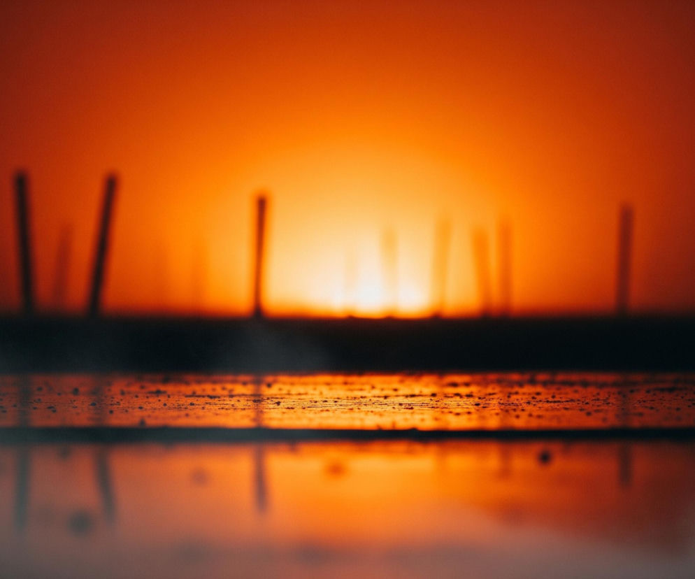 Pomarańczowe słońce, intensywnie świecące, dominuje tło obrazu, jego blask rozlewa się na cały kadr, tworząc gradient od jasnej żółci w centrum do głębokiego pomarańczu na brzegach. Na horyzoncie widoczne są ciemne, smukłe sylwetki obiektów przypominających słupy lub kominy, rozrzucone w nieregularnych odstępach, a ich kształty są rozmyte i niewyraźne. Pierwszy plan przedstawia ciemną, poziomą powierzchnię pokrytą licznymi drobnymi kropelkami, które odbijają pomarańczowy blask, zaś pod nią widoczne jest rozmyte odbicie sceny, z ciemnymi plamami przypominającymi słupy i fragmentami mgły unoszącej się przy dolnych krawędziach kadru.